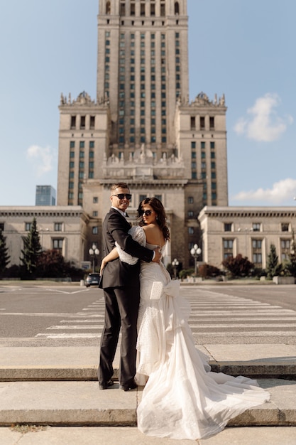 Jungvermählten, Mann und Frau im Hochzeitskleid gehen entlang des Fußgängerüberwegs vor dem Hintergrund der Stadtlandschaft, drehen sich um Lächeln, glücklich zusammen, Konzept des Hochzeitstages. Glückliche Momente