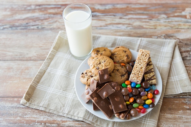 Junk Food, Süßigkeiten und ungesundes Esskonzept - Nahaufnahme von Süßigkeiten, Schokolade, Müsli und Keksen mit Milchglas auf dem Teller