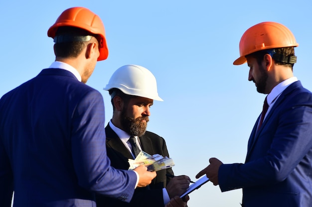 La junta de arquitectos con rostros interesados en trajes y cascos toma notas y retiene dinero. Concepto de construcción y trato. Los trabajadores y el ingeniero celebran una reunión sobre el fondo del cielo azul. Los constructores hacen gangas