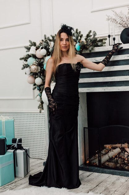 Foto junto a la chimenea de año nuevo adornado posando a jovencita en vestido largo negro