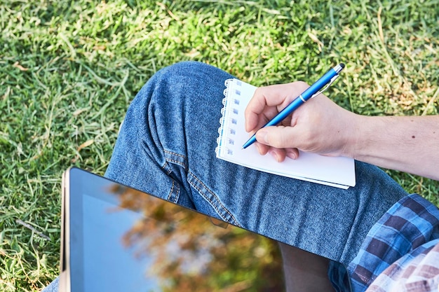 Foto junto a un joven irreconocible escribiendo en un cuaderno usando un bolígrafo estudiando o trabajando al aire libre