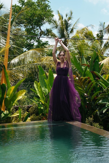 Foto junto a la piscina, a su lado, está de pie con un vestido de noche de chica contra un fondo de palmeras.