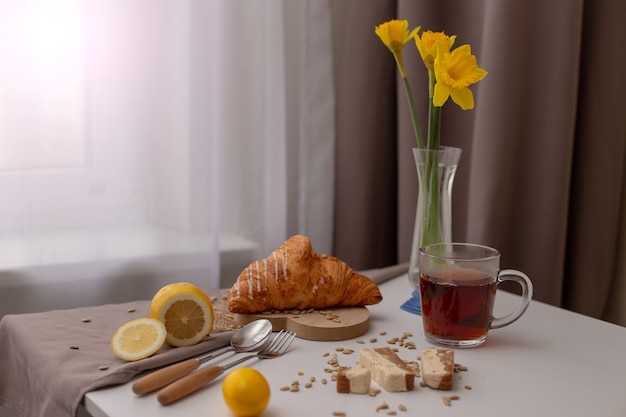 Junto a la ventana con cortinas blancas y marrones, sobre una mesa blanca, se sirve el desayuno con una taza de té, croissants, limones y narcisos amarillos en un florero de cristal.