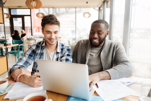 Juntos es más eficiente. Dos chicos guapos felices están usando una computadora portátil moderna en la cafetería mientras estudian.