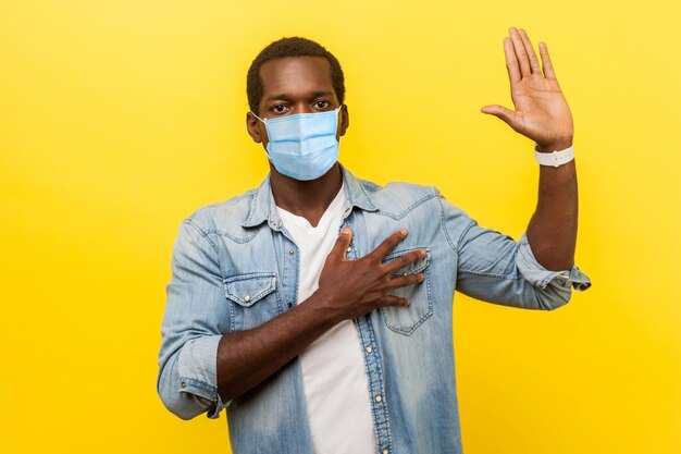 Juro. Retrato de homem sério honesto em camisa casual jeans com máscara médica cirúrgica, mantendo a mão no peito, levantando a palma da mão, dando promessa, promessa. tiro de estúdio interno isolado em fundo amarelo