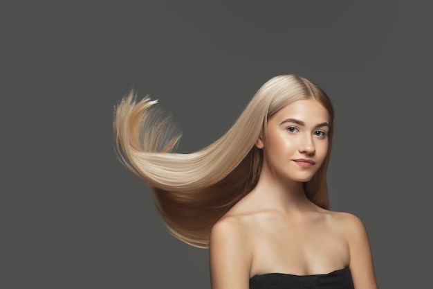 Juventud. Hermosa modelo con cabello rubio largo, liso y volador sobre fondo gris oscuro de estudio. Modelo caucásico joven con piel bien cuidada y cabello soplando en el aire. Concepto de cuidado de salón, belleza, moda.