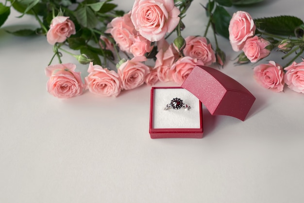 Juwelenbesetzter Verlobungsring in geöffneter roter Schmuckschatulle, umgeben von blassrosa Rosen. Valentinstag feiern