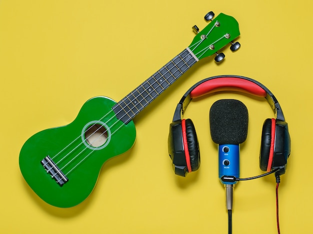 Kabelgebundener Kopfhörer blau verkabelte MIC und Gitarrenukulele grün auf gelbem Grund. Ausrüstung zum Aufnehmen von Musiktiteln. Der Blick von oben. Flach liegen.