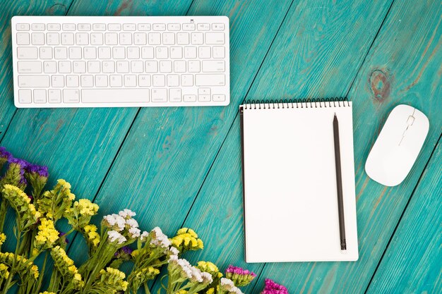 Kabelloser, schlanker Tastatur-Maus-Notizblock mit Bleistift und bunten Blumen auf blauem Holztisch