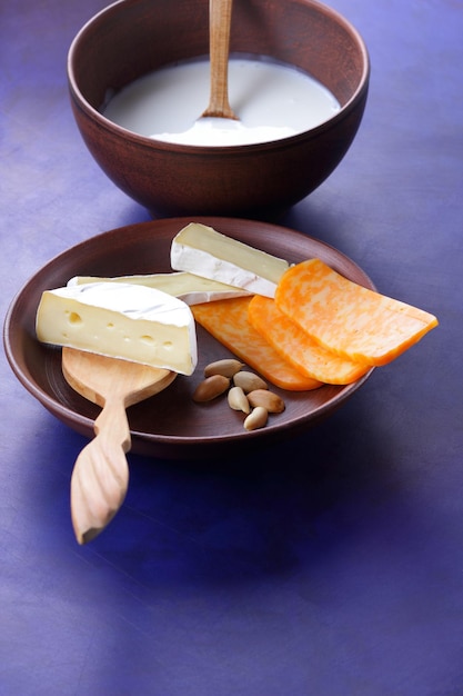 Käse mit Nüssen und einem Holzspatel auf einer Tonplatte Verschiedene Käsesorten und eine Schüssel Milch auf blauem Hintergrund, Nahaufnahme