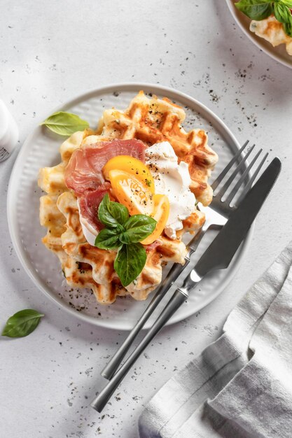 Käse- oder Kartoffelwaffeln, serviert mit gelben Tomaten, Frischkäse, Schinken und Basilikumblättern mit Besteck Leckeres, herzhaftes Frühstück Draufsicht flach gelegt