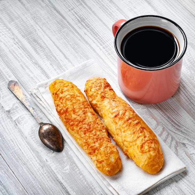 Käsebrötchen, Brot mit Käse und eine Tasse Kaffee mit einem Löffel auf weißem Holzhintergrund