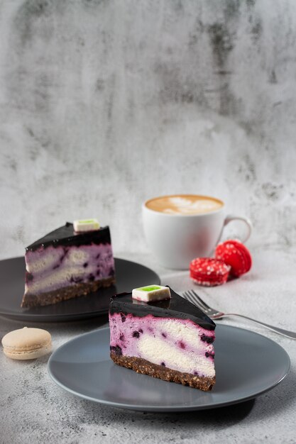 Käsekuchen mit Blaubeere mit Tasse Kaffee auf weißem Tisch. Nahaufnahme. Leckeres Frühstück. Stück Kuchen auf schwarzem Teller, weiße Tasse auf weißem Marmorhintergrund. Vertikales Foto.