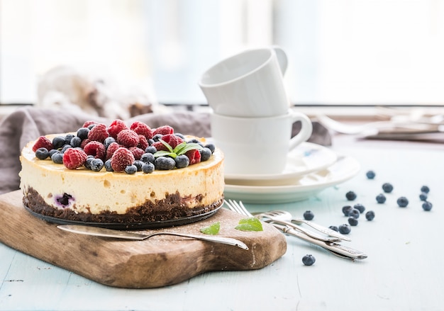 Käsekuchen mit frischen Himbeeren und Blaubeeren auf einem hölzernen Umhüllungsbrett, Platten, Schalen, Küchenserviette, Tafelsilber über blauem Hintergrund, Fenster am Hintergrund.