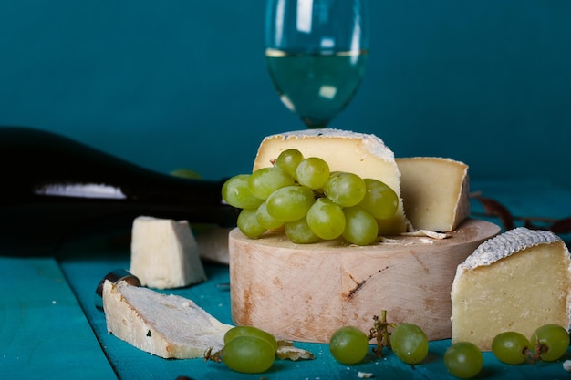 Käsescheiben, weiße Trauben, Messer, Glas und eine Flasche Weißwein auf einer Holzoberfläche. Nahaufnahme