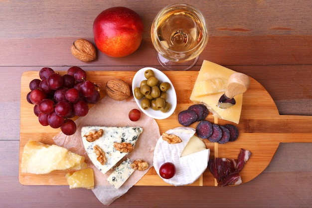 Käsesortiment mit Früchten, Trauben, Nüssen, Glas mit Wein und Käsemesser auf einem hölzernen Serviertablett