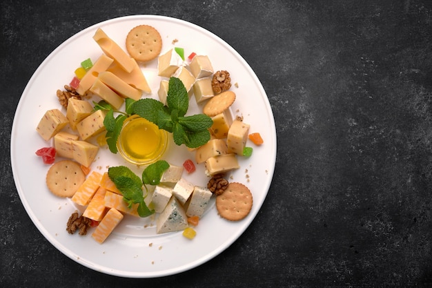 Käseteller, verschiedene Käsesorten mit Minze, kandierten Früchten, Honig und Keksen, auf einem weißen Teller, auf dunklem Hintergrund