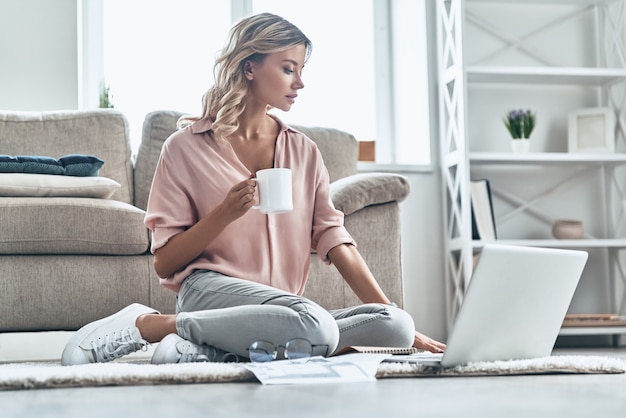 Kaffee bringt frische Ideen. Schöne junge Frau, die eine Tasse hält und einen Laptop benutzt, während sie zu Hause auf dem Boden sitzt