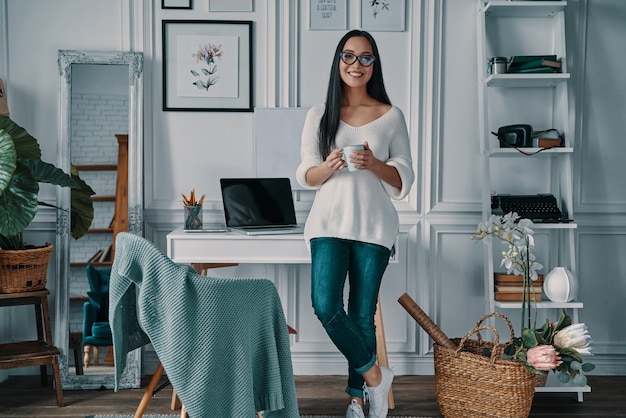 Kaffee bringt frische Ideen. Schöne junge Frau, die in die Kamera schaut und lächelt, während sie im Home Office steht