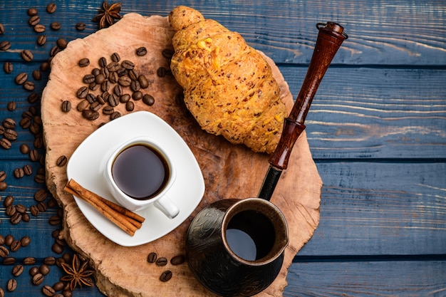 Kaffee gießt in eine Tasse auf einem blauen Holztisch mit Kaffeebohnen und einem Croissant.