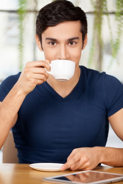 Kaffee im Restaurant genießen. Schöner junger Mann, der Kaffee im Restaurant trinkt und lächelt, während er ein digitales Tablet auf dem Tisch legt