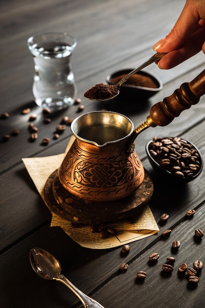 Kaffee in Cezve zubereiten. Bohnen, Hand mit einem Löffel gemahlenen Kaffee, Cezve, Wasser in einem Glas. Hölzerner Hintergrund.