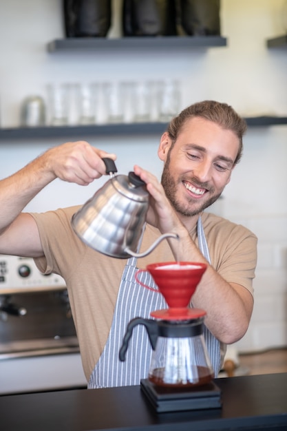 Kaffee kochen. lächelnder mann in einer schürze, die im café steht und wasser in übergießen von der teekanne gießt, die kaffee macht