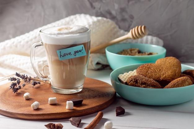 Kaffee Latte Tasse mit Liebe Sie Aufkleber auf Holztablett mit türkisfarbenen Schalen mit Keksen und Marshmallows auf grauer Wandoberfläche