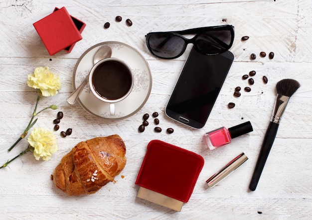 Kaffee mit Croissant, Handy, Sonnenbrille und Make-up-Tools auf einem Tisch