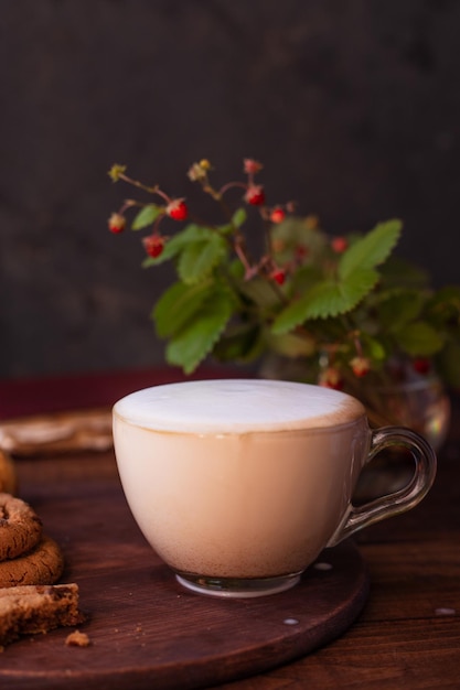 Kaffee mit Milchschaum in einem Glasbecher auf einem Holztisch neben einem Blumenstrauß aus Wildblumen und Keksen