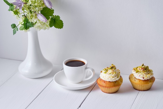 Kaffee- und Obst-Cupcakes zum Frühstück. Morgentisch mit Dessert, Espresso und Blumen in einer Vase auf einem weißen Holztisch.