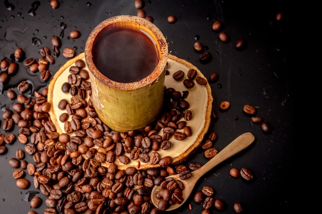 Kaffeebecher aus Bambus auf Holzuntertasse und viele geröstete Kaffeebohnen auf schwarzem Hintergrund verstreut, dunkler Kaffee hat Rauch und Aroma, Getränk für die Pause, Morgen- und Kaffeezeit, lokaler Stil.