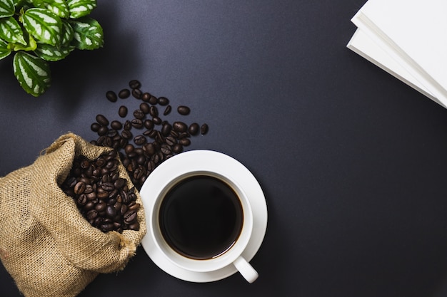 Kaffeebohnen in einer Sacktasche und in einem schwarzen Kaffee in einer weißen Kaffeetasse, Bäume, Bücher auf einem schwarzen Hintergrund