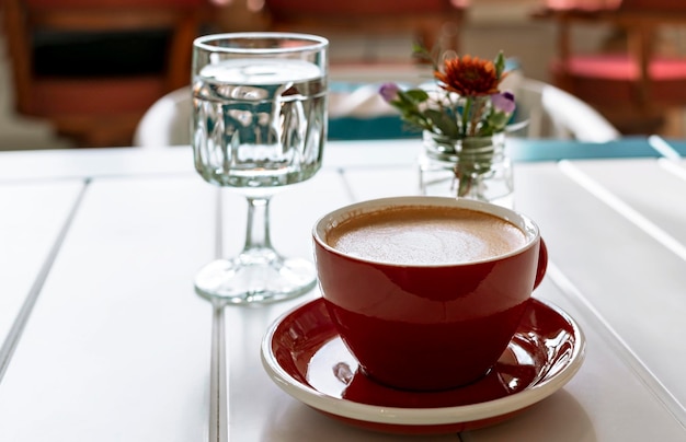 Kaffeecappuccino im roten Schalenglas Wasser und kleine Blumensträuße im Glasgefäß auf weißem Tisch