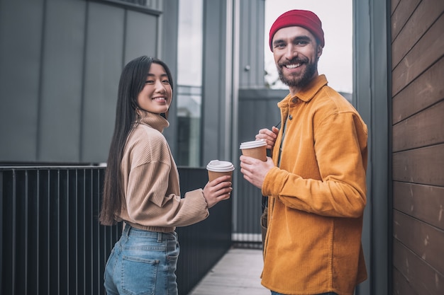 Kaffeepause. Junger Mann und junge Frau in der Freizeitkleidung, die beim Kaffee draußen positiv schaut