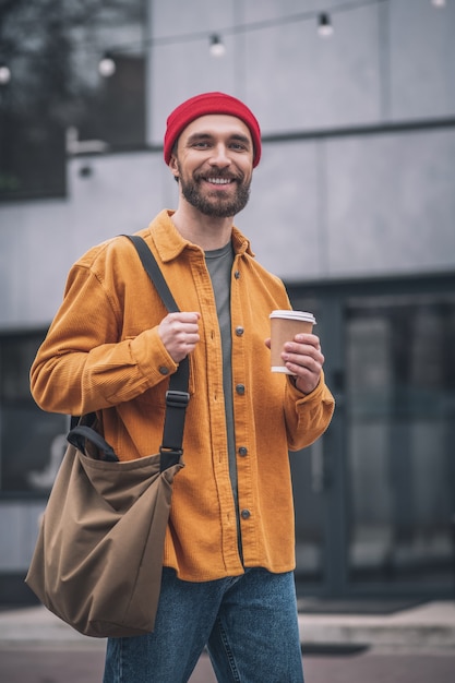 Kaffeepause. Mann in einem roten Hut und in einer orangefarbenen Jacke mit einer Kaffeetasse in den Händen