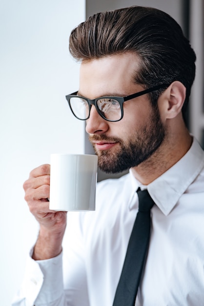 Kaffeepause. Nachdenklicher junger Geschäftsmann in Gläsern, die Kaffee trinken und wegschauen, während sie im Büro stehen