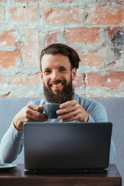 Kaffeepause und Arbeit. Mann hält eine Tasse heißes Getränk vor Laptop sitzen.