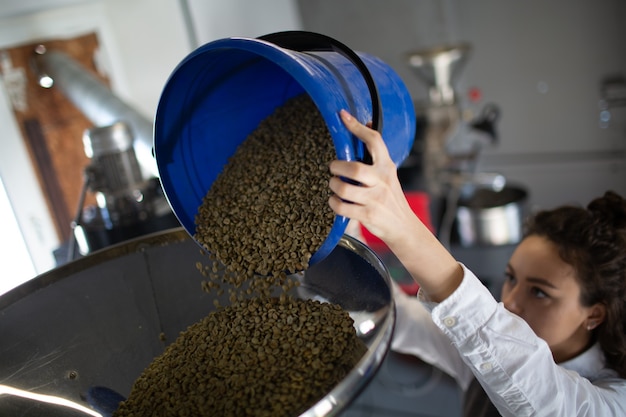 Kaffeeröstermaschine beim Kaffeerösten. Junge Arbeiterin Barista, die grüne Kaffeebohnen in Apparat lädt.