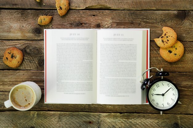 Kaffeetasse, geöffnetes Buch, Wecker, Plätzchen auf hölzerner rustikaler Weinlese.