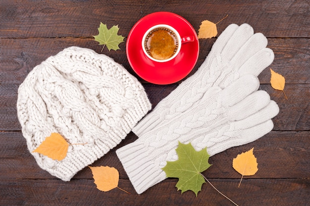 Kaffeetasse, Handschuhe, handgemachte Strickmütze und Herbstlaub auf hölzernen Brettern