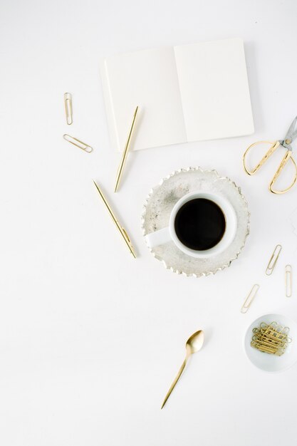 Kaffeetasse, leeres Tagebuch und goldenes Zubehör: Teelöffel, Stift, Clips und Schere auf Weiß