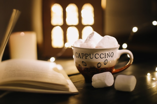 Kaffeetasse mit Marshmallows und einem Buch auf dem Tisch. Stillleben auf dunklem Hintergrund, flach lag