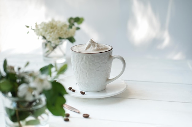 Kaffeetasse mit Schlagsahne auf einem weißen Holztisch, Cappuccino in einer weißen Tasse, trendiges Morgenlicht mit Blumen und Kaffeebohnen, Guten-Morgen-Konzept