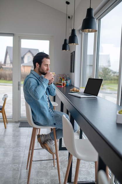 Kaffeezeit. Junger bärtiger Mann, der Kaffee trinkt, sitzt seitlich zur Kamera am Tisch mit Laptop drinnen in der Nähe der Glaswand