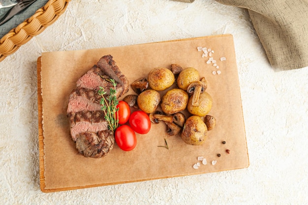 Kalbssteak mit Ofenkartoffeln und Champignons. Mit Kräutern und Kirschtomaten dekoriert. Auf einem Holzbrett serviert. Sicht von oben.