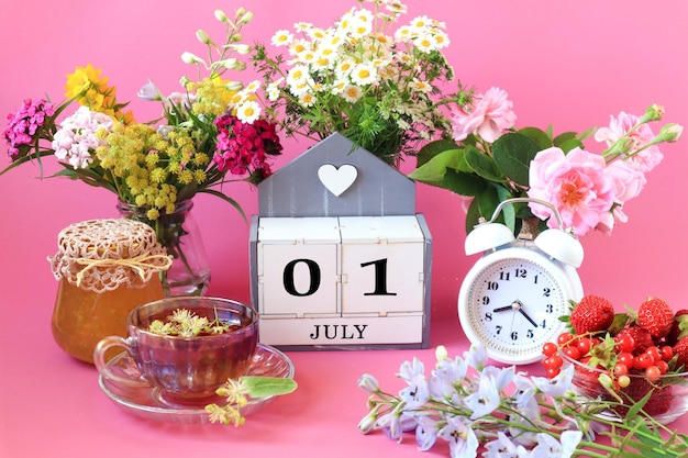 Kalender für den 1. Juli der Name des Monats Juli in englischen Würfeln mit den Zahlen 0 und 1 eine Tasse Tee Blumensträuße ein Wecker Beeren auf rosa Hintergrund