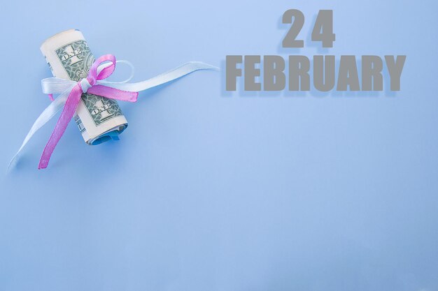 Kalenderdatum auf blauem Hintergrund mit aufgerollten Dollarscheinen, die von blauem und rosafarbenem Band mit Kopierraum festgehalten werden. Der 24. Februar ist der vierundzwanzigste Tag des Monats.