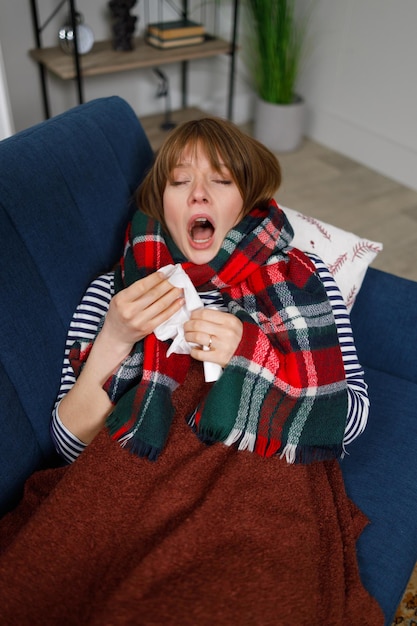 Kalte Frau, die zu Hause auf dem Sofa liegt, niest in ein Papiertaschentuch