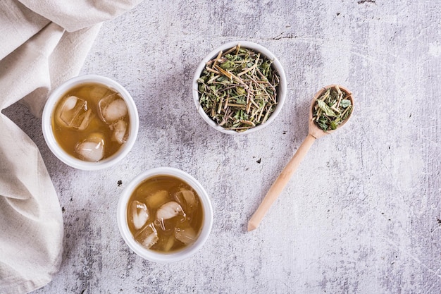 Kalter grüner japanischer Hojicha-Tee mit Eis in Tassen und trockener Tee in einer Schüssel auf dem Tisch Draufsicht